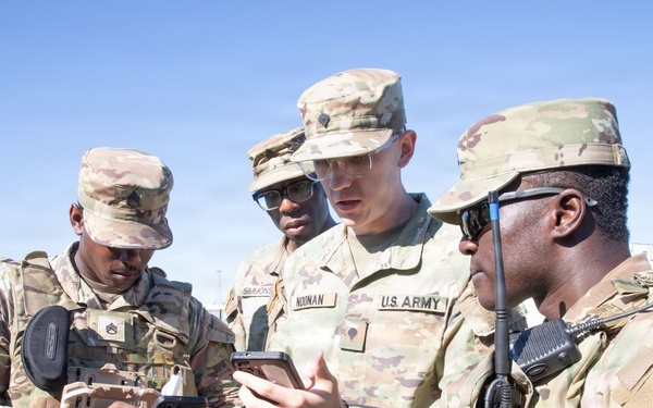 Four U.S. Army soldiers in OCP (Operational Camouflage Pattern) uniforms are gathered outdoors, reviewing information on mobile devices. The soldiers appear to be coordinating or sharing information, with at least two holding smartphones and one equipped with a radio. The setting appears to be a military installation or training area with a clear blue sky and green grass visible in the background.