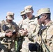 Four U.S. Army soldiers in OCP (Operational Camouflage Pattern) uniforms are gathered outdoors, reviewing information on mobile devices. The soldiers appear to be coordinating or sharing information, with at least two holding smartphones and one equipped with a radio. The setting appears to be a military installation or training area with a clear blue sky and green grass visible in the background.