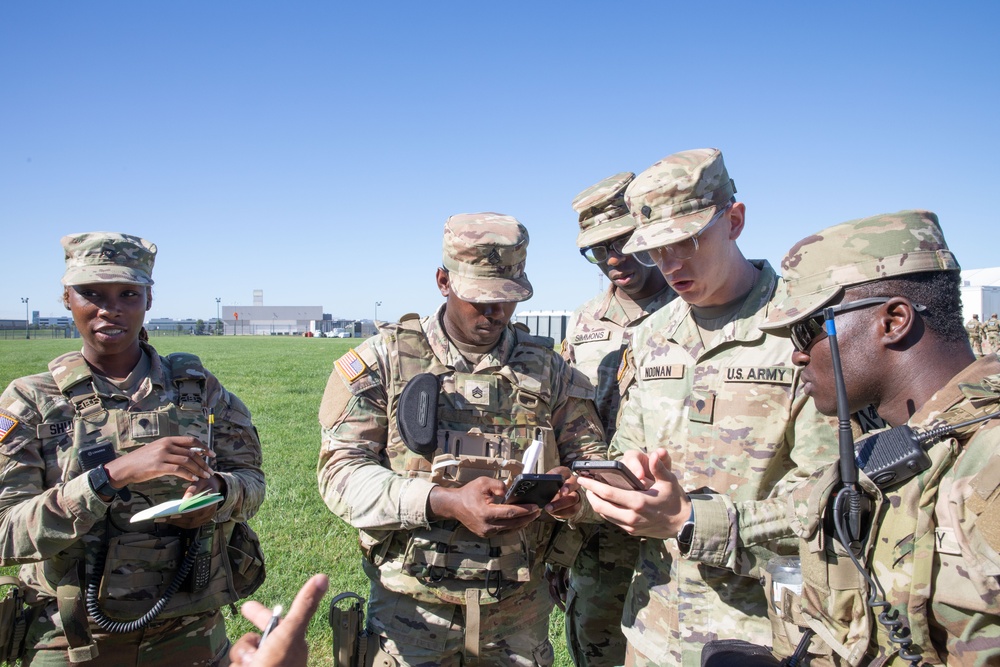 A group of U.S. Army soldiers in OCP (Operational Camouflage Pattern) uniforms are gathered outdoors on a grassy field, reviewing information on a mobile device. The soldiers appear to be in a coordination or briefing activity, with visible name tapes including 'SIMMONS,' 'NOONAN,' and partially visible others. The setting appears to be a military installation or airfield based on the background structures.