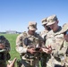 A group of U.S. Army soldiers in OCP (Operational Camouflage Pattern) uniforms are gathered outdoors on a grassy field, reviewing information on a mobile device. The soldiers appear to be in a coordination or briefing activity, with visible name tapes including 'SIMMONS,' 'NOONAN,' and partially visible others. The setting appears to be a military installation or airfield based on the background structures.