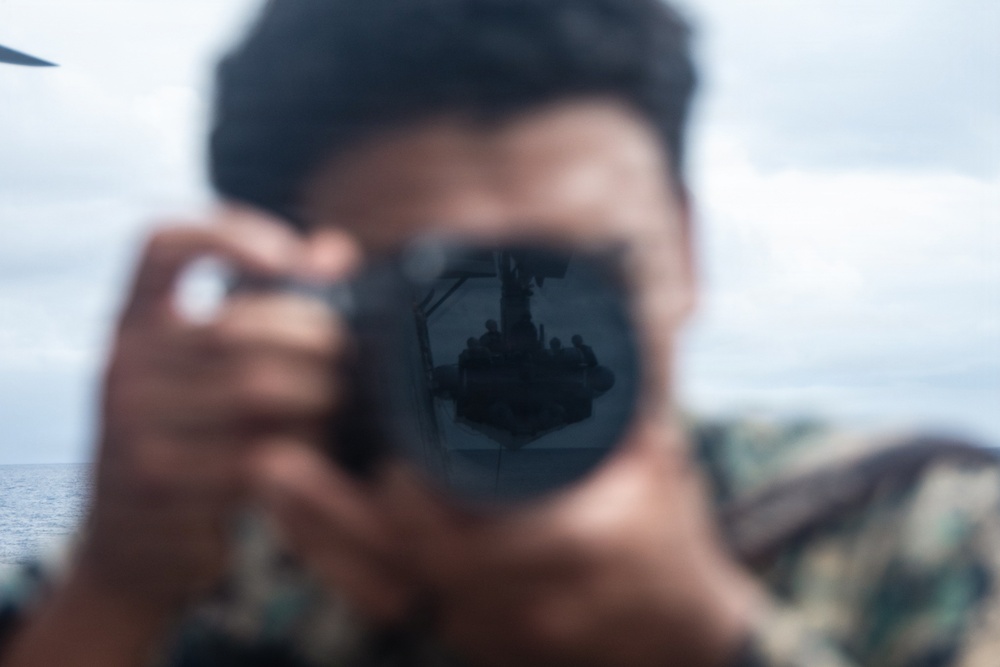 A person wearing camouflage military uniform holds a camera directly toward the viewer, with the lens in sharp focus while the subject remains blurred. Reflected in the camera lens is a scene showing what appears to be a naval vessel deck with personnel and equipment visible. The image is taken in an outdoor maritime environment with overcast skies and open water visible in the background.