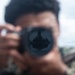 A person wearing camouflage military uniform holds a camera directly toward the viewer, with the lens in sharp focus while the subject remains blurred. Reflected in the camera lens is a scene showing what appears to be a naval vessel deck with personnel and equipment visible. The image is taken in an outdoor maritime environment with overcast skies and open water visible in the background.