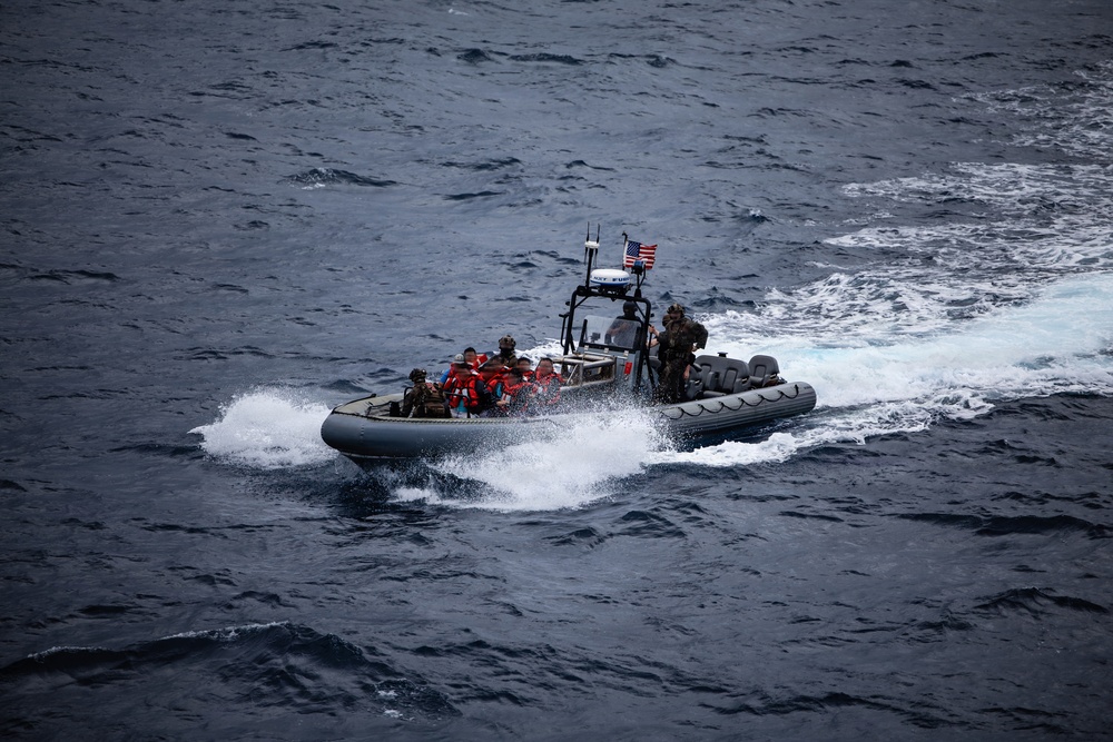 A rigid-hull inflatable boat (RHIB) is underway at speed on open ocean waters. The vessel carries multiple personnel, some wearing orange life vests and others in military/tactical gear, suggesting a rescue or boarding operation. A United States flag is visibly mounted on the boat's console structure.