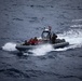 A rigid-hull inflatable boat (RHIB) is underway at speed on open ocean waters. The vessel carries multiple personnel, some wearing orange life vests and others in military/tactical gear, suggesting a rescue or boarding operation. A United States flag is visibly mounted on the boat's console structure.