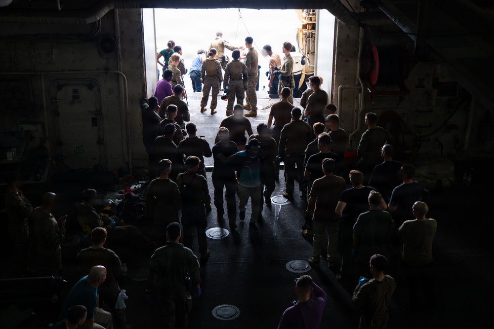 A large group of military personnel, likely Marines based on their uniforms and haircuts, are gathered inside what appears to be the well deck or cargo bay of a naval vessel. They are oriented toward a large open hatch or ramp at the rear of the ship, through which bright daylight is visible. A smaller group near the opening appears to be receiving instructions or conducting a briefing.