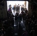 A large group of military personnel, likely Marines based on their uniforms and haircuts, are gathered inside what appears to be the well deck or cargo bay of a naval vessel. They are oriented toward a large open hatch or ramp at the rear of the ship, through which bright daylight is visible. A smaller group near the opening appears to be receiving instructions or conducting a briefing.