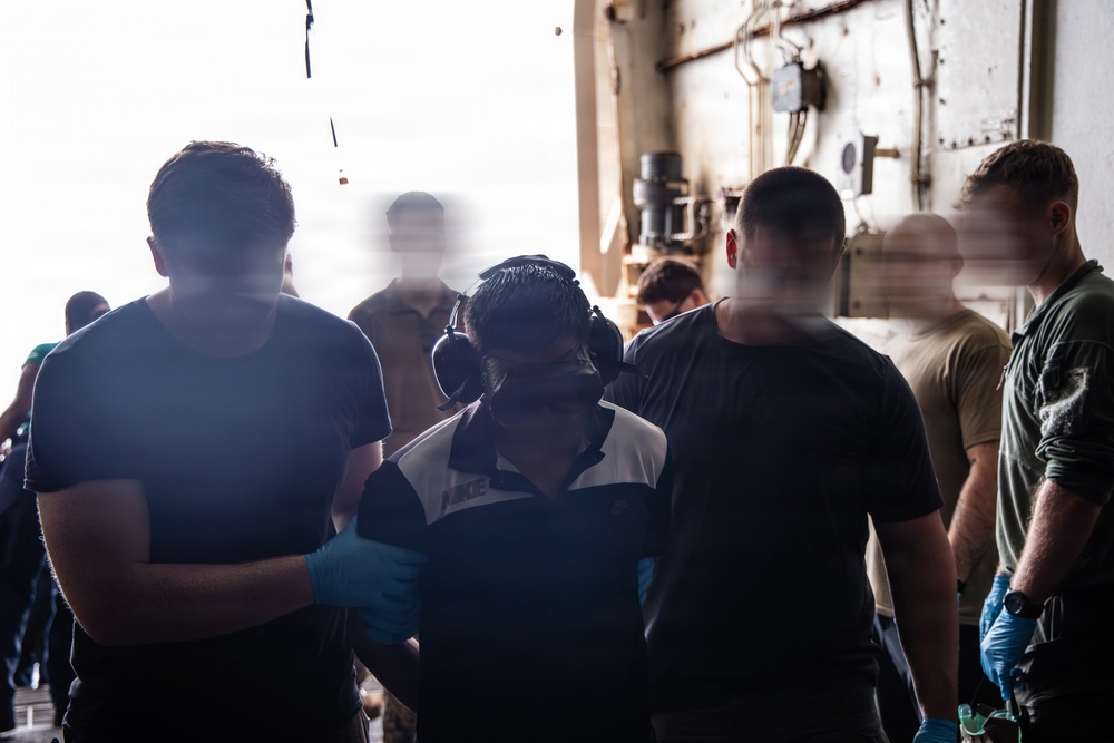 A group of individuals are gathered in what appears to be an industrial or military vessel interior space. Several people are wearing casual clothing including Nike apparel, with at least one person wearing hearing protection (ear muffs) and blue nitrile gloves. The scene suggests a work or training activity taking place in a confined, utilitarian environment.