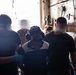 A group of individuals are gathered in what appears to be an industrial or military vessel interior space. Several people are wearing casual clothing including Nike apparel, with at least one person wearing hearing protection (ear muffs) and blue nitrile gloves. The scene suggests a work or training activity taking place in a confined, utilitarian environment.