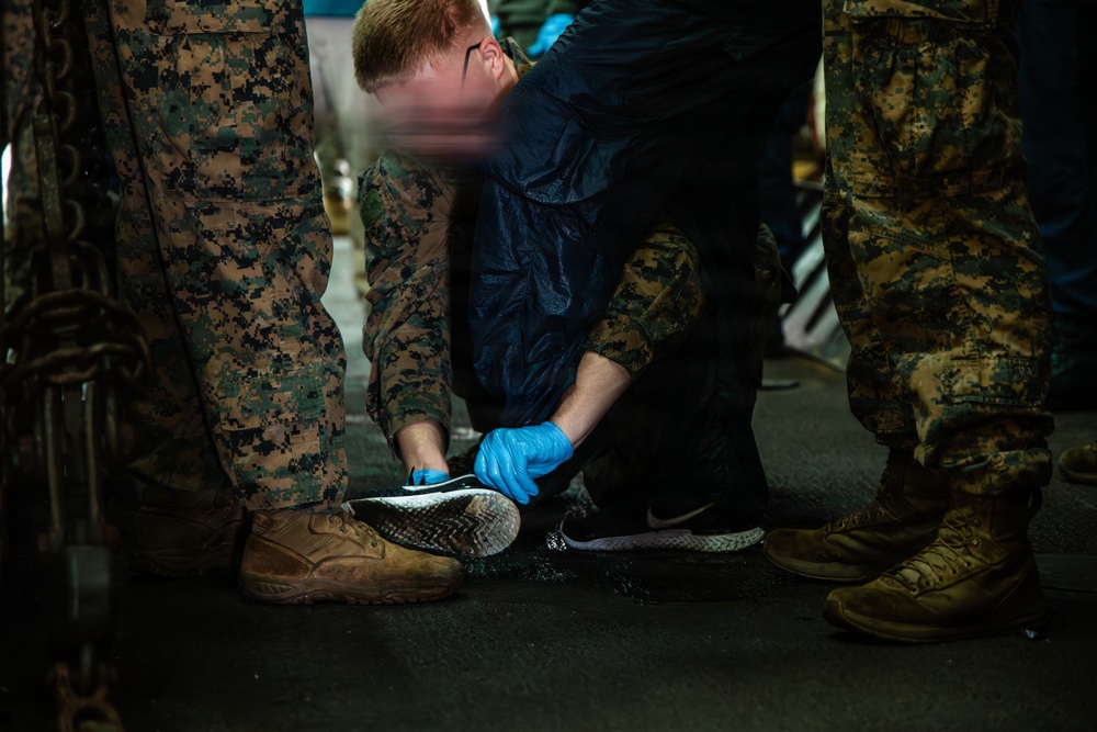 A person wearing blue latex gloves and a dark rain jacket is crouched down cleaning or inspecting the sole of an athletic shoe (Nike brand) on a wet surface. They are surrounded by individuals in U.S. Marine Corps MARPAT digital camouflage uniforms and tan combat boots. The scene appears to take place in a dimly lit, industrial or military environment.