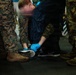 A person wearing blue latex gloves and a dark rain jacket is crouched down cleaning or inspecting the sole of an athletic shoe (Nike brand) on a wet surface. They are surrounded by individuals in U.S. Marine Corps MARPAT digital camouflage uniforms and tan combat boots. The scene appears to take place in a dimly lit, industrial or military environment.