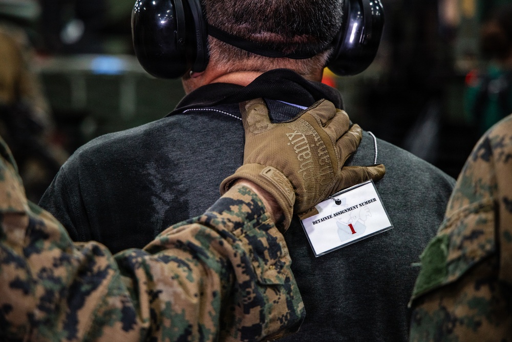 A military personnel wearing MARPAT (Marine digital camouflage) uniform and tan Mechanix tactical gloves has a hand placed on the back of an individual wearing civilian clothing and hearing protection. The individual has a badge attached to their back reading 'DETAINEE ASSIGNMENT NUMBER 1', indicating this is a military training exercise involving detainee handling procedures. The scene appears to be inside a military vehicle or facility.