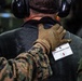 A military personnel wearing MARPAT (Marine digital camouflage) uniform and tan Mechanix tactical gloves has a hand placed on the back of an individual wearing civilian clothing and hearing protection. The individual has a badge attached to their back reading 'DETAINEE ASSIGNMENT NUMBER 1', indicating this is a military training exercise involving detainee handling procedures. The scene appears to be inside a military vehicle or facility.