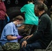 A medical professional in blue scrubs and military camouflage pants, wearing a surgical mask and purple gloves, is attending to a seated civilian male patient while writing on a clipboard. The scene appears to be a field medical treatment area, likely a military humanitarian or disaster relief operation. Multiple personnel in various uniforms and scrubs are visible in the background, along with a large military vehicle tire.