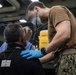 A military medical personnel wearing U.S. Navy/Marine Corps digital camouflage (MARPAT) trousers and a tan t-shirt is using a stethoscope to examine a seated individual wearing a black shirt with a badge labeled 'DETAINEE ASSIGNMENT NUMBER'. The medical provider is wearing blue nitrile gloves and a surgical face mask, suggesting a clinical or field medical screening context. A yellow sharps disposal container is visible in the background.