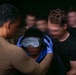 A person wearing blue nitrile gloves and an olive/tan shirt is fitting a helmet or headgear device with taped-over goggles onto another individual. The scene appears to be a training or demonstration exercise, with several other individuals visible in the background. The goggles appear to have been deliberately obscured with tape, suggesting a sensory deprivation or disorientation training scenario.