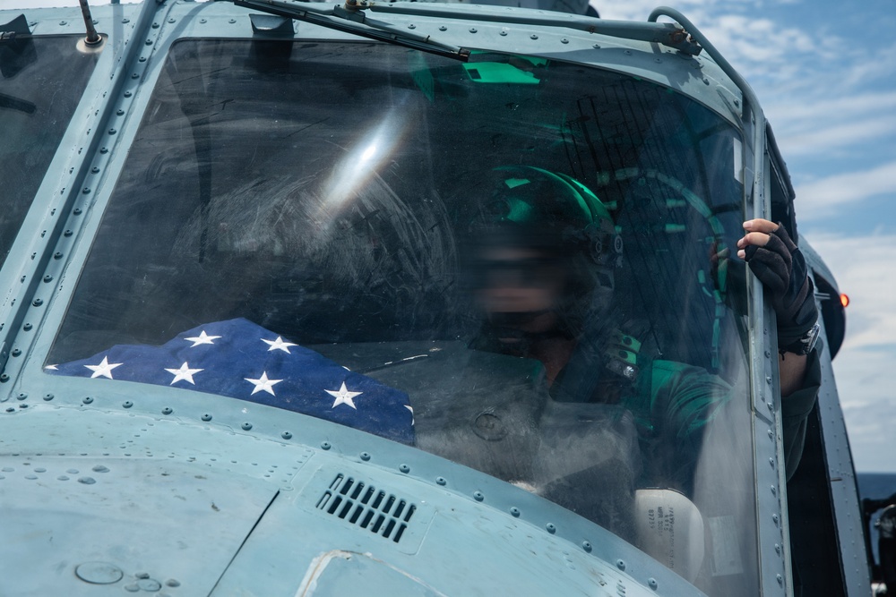 A military helicopter cockpit is shown with a pilot or crew member wearing a flight helmet and oxygen mask seated inside. A folded American flag with visible white stars on a blue field is prominently placed in the cockpit. A second crew member's hand is visible gripping the cockpit frame from outside.
