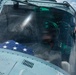 A military helicopter cockpit is shown with a pilot or crew member wearing a flight helmet and oxygen mask seated inside. A folded American flag with visible white stars on a blue field is prominently placed in the cockpit. A second crew member's hand is visible gripping the cockpit frame from outside.