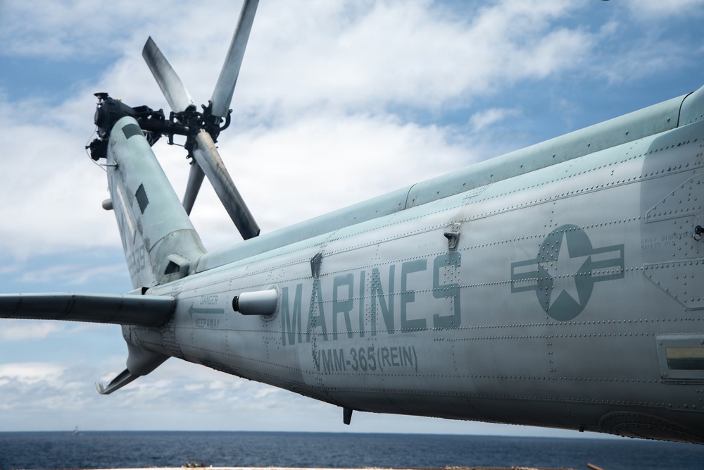 A close-up photograph of a U.S. Marines military helicopter, specifically belonging to VMM-365 (REIN), a Marine Medium Tiltrotor Squadron. The aircraft is painted in a light gray color scheme and features the USMC roundel insignia alongside bold 'MARINES' lettering on the fuselage. The helicopter appears to be parked on a naval vessel deck with ocean and cloudy sky visible in the background.