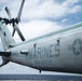 A close-up photograph of a U.S. Marines military helicopter, specifically belonging to VMM-365 (REIN), a Marine Medium Tiltrotor Squadron. The aircraft is painted in a light gray color scheme and features the USMC roundel insignia alongside bold 'MARINES' lettering on the fuselage. The helicopter appears to be parked on a naval vessel deck with ocean and cloudy sky visible in the background.