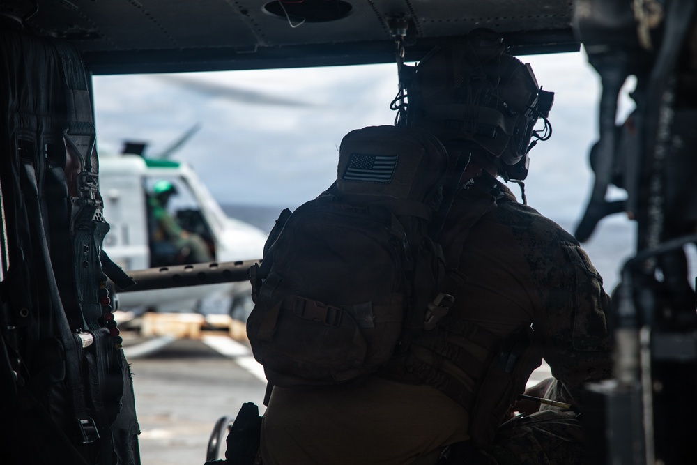 A U.S. military personnel, identifiable by the American flag patch on their shoulder, is seated inside a helicopter looking out toward another helicopter on what appears to be a naval vessel flight deck. The individual is wearing full tactical gear including a helmet with attachments, body armor, and a backpack. The scene suggests a maritime military operation or exercise.
