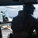 A U.S. military personnel, identifiable by the American flag patch on their shoulder, is seated inside a helicopter looking out toward another helicopter on what appears to be a naval vessel flight deck. The individual is wearing full tactical gear including a helmet with attachments, body armor, and a backpack. The scene suggests a maritime military operation or exercise.