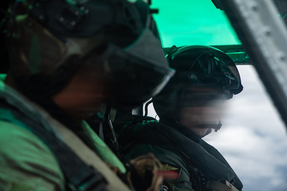 Two military aviators are seated inside a cockpit of a military aircraft in flight. Both crew members are wearing flight helmets with visors and appear to be on an active mission, with clouds visible through the cockpit window. A green-tinted heads-up display or canopy tint is visible in the upper portion of the frame.