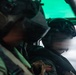 Two military aviators are seated inside a cockpit of a military aircraft in flight. Both crew members are wearing flight helmets with visors and appear to be on an active mission, with clouds visible through the cockpit window. A green-tinted heads-up display or canopy tint is visible in the upper portion of the frame.