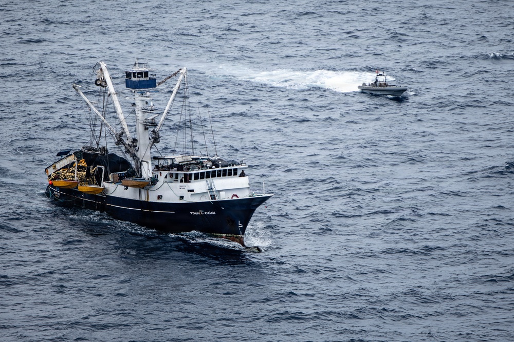 An aerial photograph shows a large commercial fishing vessel with visible fire or burn damage on its deck, accompanied by a smaller patrol or response boat nearby. The fishing vessel appears to be stationary or moving slowly in open ocean waters, while the smaller boat is actively underway, leaving a visible wake. The scene suggests an emergency response or law enforcement interaction at sea.