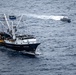 An aerial photograph shows a large commercial fishing vessel with visible fire or burn damage on its deck, accompanied by a smaller patrol or response boat nearby. The fishing vessel appears to be stationary or moving slowly in open ocean waters, while the smaller boat is actively underway, leaving a visible wake. The scene suggests an emergency response or law enforcement interaction at sea.