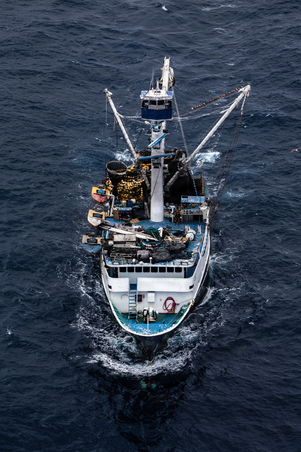 An aerial photograph taken from directly above showing a commercial fishing vessel underway on open dark blue ocean. The vessel appears to be a purse seiner or trawler, heavily laden with fishing gear, nets, and buoys on its working deck. The ship is moving forward, creating a visible bow wake and stern wake.