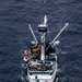 An aerial photograph taken from directly above showing a commercial fishing vessel underway on open dark blue ocean. The vessel appears to be a purse seiner or trawler, heavily laden with fishing gear, nets, and buoys on its working deck. The ship is moving forward, creating a visible bow wake and stern wake.