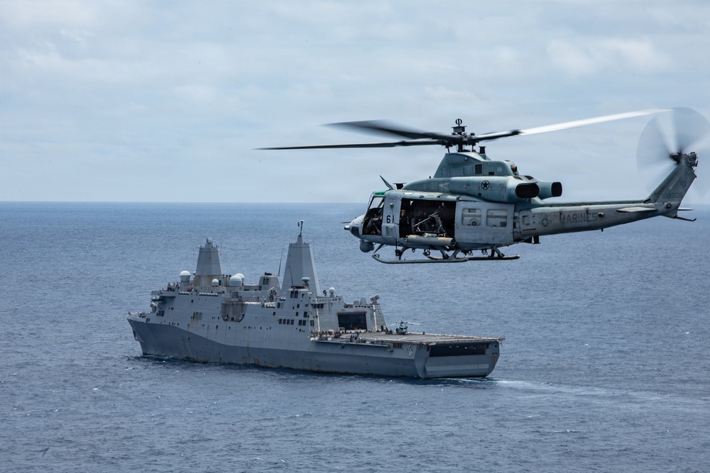A U.S. Marines UH-1Y Venom helicopter flies in close proximity to a U.S. Navy San Antonio-class amphibious transport dock ship over open ocean. The helicopter bears the number '61' and 'MARINES' markings, with its side door open and rotor blades in motion. The scene depicts a maritime military operation, likely a training exercise or operational deployment.