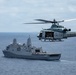 A U.S. Marines UH-1Y Venom helicopter flies in close proximity to a U.S. Navy San Antonio-class amphibious transport dock ship over open ocean. The helicopter bears the number '61' and 'MARINES' markings, with its side door open and rotor blades in motion. The scene depicts a maritime military operation, likely a training exercise or operational deployment.