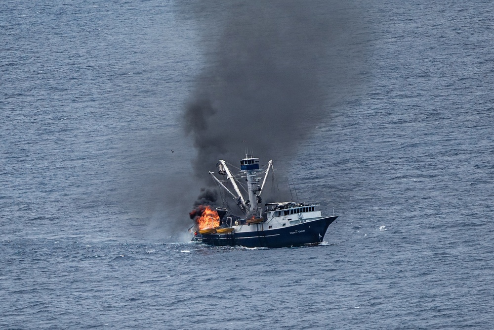 A commercial fishing vessel is on fire in open ocean waters, with large orange flames visible on the forward/mid section of the boat and a thick column of black smoke rising high into the sky. The vessel appears to be a trawler or seiner type fishing boat with outrigger booms and a wheelhouse still intact. The image was taken from an aerial perspective, likely from a Coast Guard or rescue aircraft.