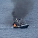A commercial fishing vessel is on fire in open ocean waters, with large orange flames visible on the forward/mid section of the boat and a thick column of black smoke rising high into the sky. The vessel appears to be a trawler or seiner type fishing boat with outrigger booms and a wheelhouse still intact. The image was taken from an aerial perspective, likely from a Coast Guard or rescue aircraft.