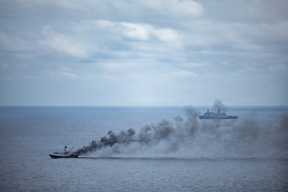 A maritime scene showing a smaller vessel on fire with heavy dark smoke billowing from it, while a larger warship is visible in the background also emitting smoke. The image appears to capture either a naval exercise, a sinking exercise (SINKEX), or an actual maritime incident at sea. The overcast sky and calm sea conditions are consistent with open ocean operations.