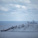 A maritime scene showing a smaller vessel on fire with heavy dark smoke billowing from it, while a larger warship is visible in the background also emitting smoke. The image appears to capture either a naval exercise, a sinking exercise (SINKEX), or an actual maritime incident at sea. The overcast sky and calm sea conditions are consistent with open ocean operations.
