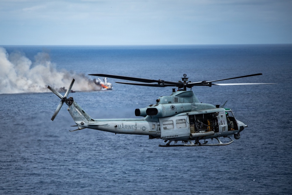 A U.S. Marines helicopter, identified as a UH-1Y Venom (serial number 168508), is flying over open ocean with its side door open and crew visible inside. In the background, a vessel appears to be on fire or producing significant smoke, suggesting a maritime exercise or incident response scenario. The image is sharp and well-composed, captured from another airborne platform.