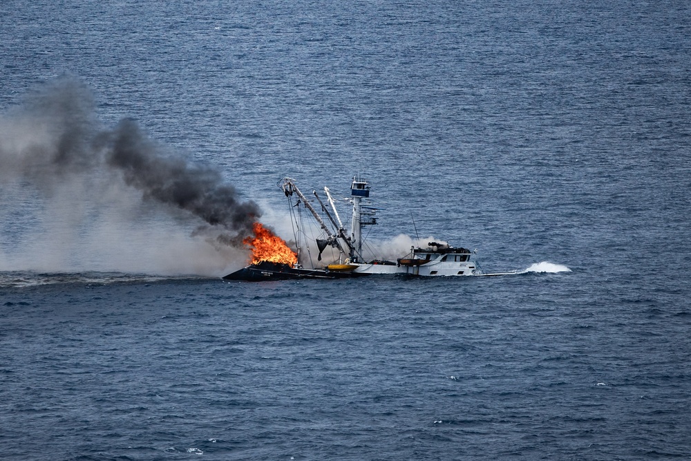 A commercial fishing vessel is engulfed in fire while underway on open ocean waters. The fire is concentrated on the forward/mid section of the vessel, producing large orange flames and a thick column of dark black smoke. The vessel appears to still be moving, suggesting engines may still be operational despite the fire.