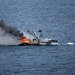 A commercial fishing vessel is engulfed in fire while underway on open ocean waters. The fire is concentrated on the forward/mid section of the vessel, producing large orange flames and a thick column of dark black smoke. The vessel appears to still be moving, suggesting engines may still be operational despite the fire.