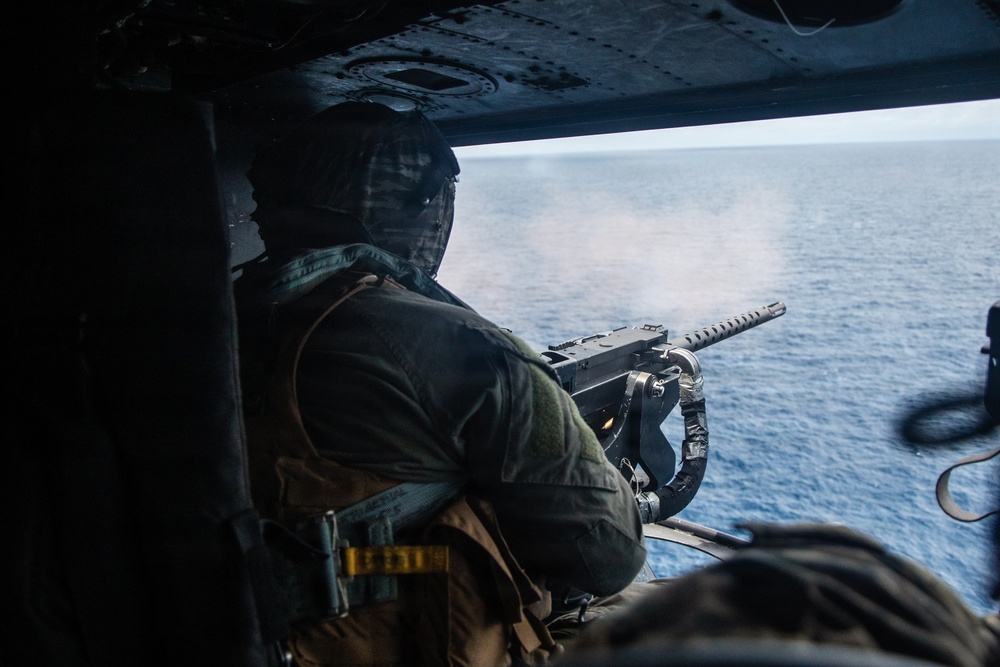 A military door gunner is firing a machine gun from the open door of a helicopter over open ocean. Smoke or gas discharge is visible at the muzzle, indicating active firing. The shooter is wearing full flight gear including a helmet with communications equipment and a tactical vest.
