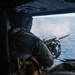 A military door gunner is firing a machine gun from the open door of a helicopter over open ocean. Smoke or gas discharge is visible at the muzzle, indicating active firing. The shooter is wearing full flight gear including a helmet with communications equipment and a tactical vest.