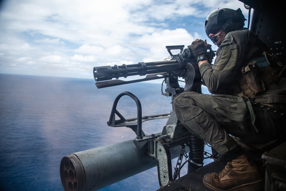 A military aircrew member is positioned at the open door of a helicopter, operating what appears to be a multi-barrel rotary cannon (likely an M134 minigun). The operator is wearing full flight gear including a helmet, flight suit, gloves, and boots, and is aiming the weapon over an open ocean. The image appears to depict a live-fire or readiness exercise conducted from a military helicopter.