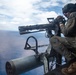 A military aircrew member is positioned at the open door of a helicopter, operating what appears to be a multi-barrel rotary cannon (likely an M134 minigun). The operator is wearing full flight gear including a helmet, flight suit, gloves, and boots, and is aiming the weapon over an open ocean. The image appears to depict a live-fire or readiness exercise conducted from a military helicopter.