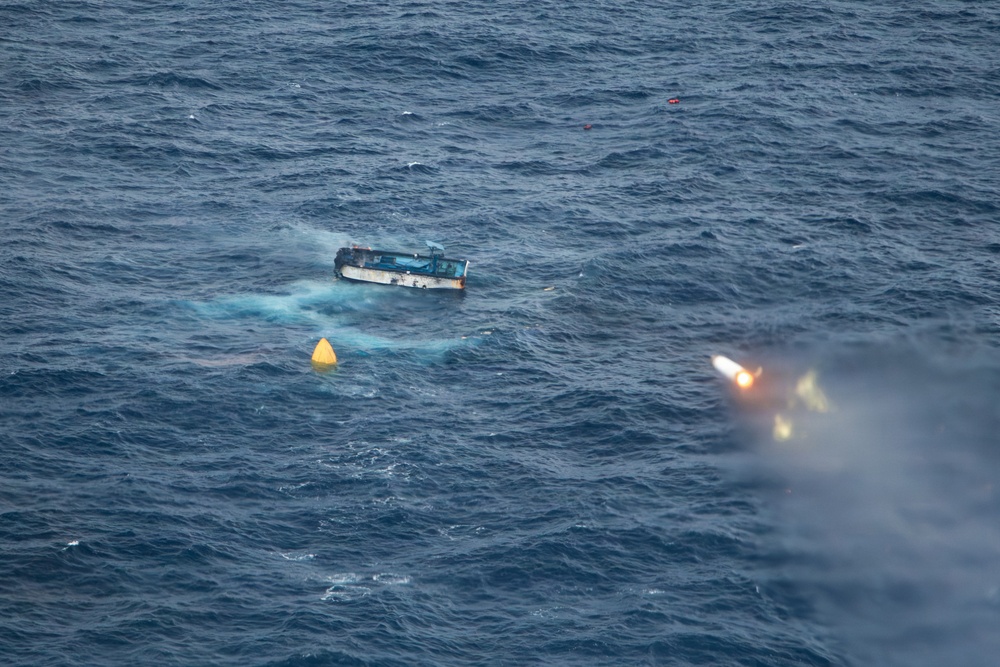 An aerial photograph shows a partially capsized or sinking small vessel in open ocean waters. A distress flare is visibly firing near the vessel, and several marker buoys are visible in the water nearby. The scene appears to depict a maritime emergency or distress situation.