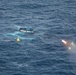 An aerial photograph shows a partially capsized or sinking small vessel in open ocean waters. A distress flare is visibly firing near the vessel, and several marker buoys are visible in the water nearby. The scene appears to depict a maritime emergency or distress situation.