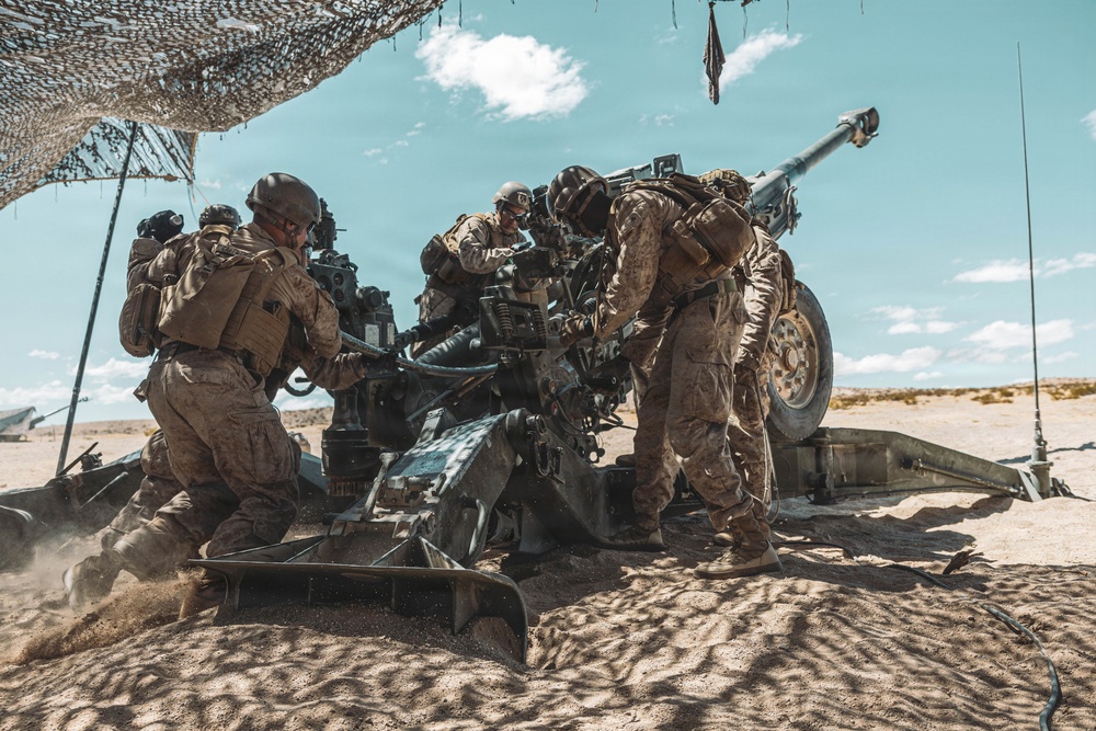 A crew of military personnel in desert camouflage uniforms are operating a large artillery piece, likely an M777 howitzer or similar towed howitzer, in an arid desert environment. The soldiers appear to be in the process of loading or servicing the weapon during what appears to be a live-fire training exercise. A camouflage netting canopy is visible overhead providing partial shade.