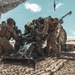 A crew of military personnel in desert camouflage uniforms are operating a large artillery piece, likely an M777 howitzer or similar towed howitzer, in an arid desert environment. The soldiers appear to be in the process of loading or servicing the weapon during what appears to be a live-fire training exercise. A camouflage netting canopy is visible overhead providing partial shade.