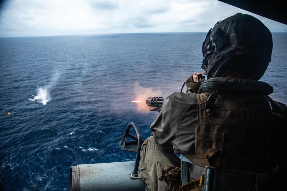 A military aircrew member is firing a multi-barrel rotary gun (consistent with an M134 minigun) from the open door of a helicopter over open ocean. Muzzle flash and smoke are visible at the weapon's barrel, and water splash impacts can be seen in the distance where rounds are striking the sea surface. The scene appears to be a live-fire training exercise conducted over open water.