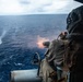 A military aircrew member is firing a multi-barrel rotary gun (consistent with an M134 minigun) from the open door of a helicopter over open ocean. Muzzle flash and smoke are visible at the weapon's barrel, and water splash impacts can be seen in the distance where rounds are striking the sea surface. The scene appears to be a live-fire training exercise conducted over open water.